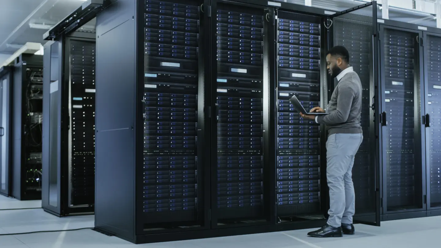 IT Specialist is Working on Laptop in Data Center while Standing Before Server Rack. Running Diagnostics, Putting in Data or Doing Maintenance Work.